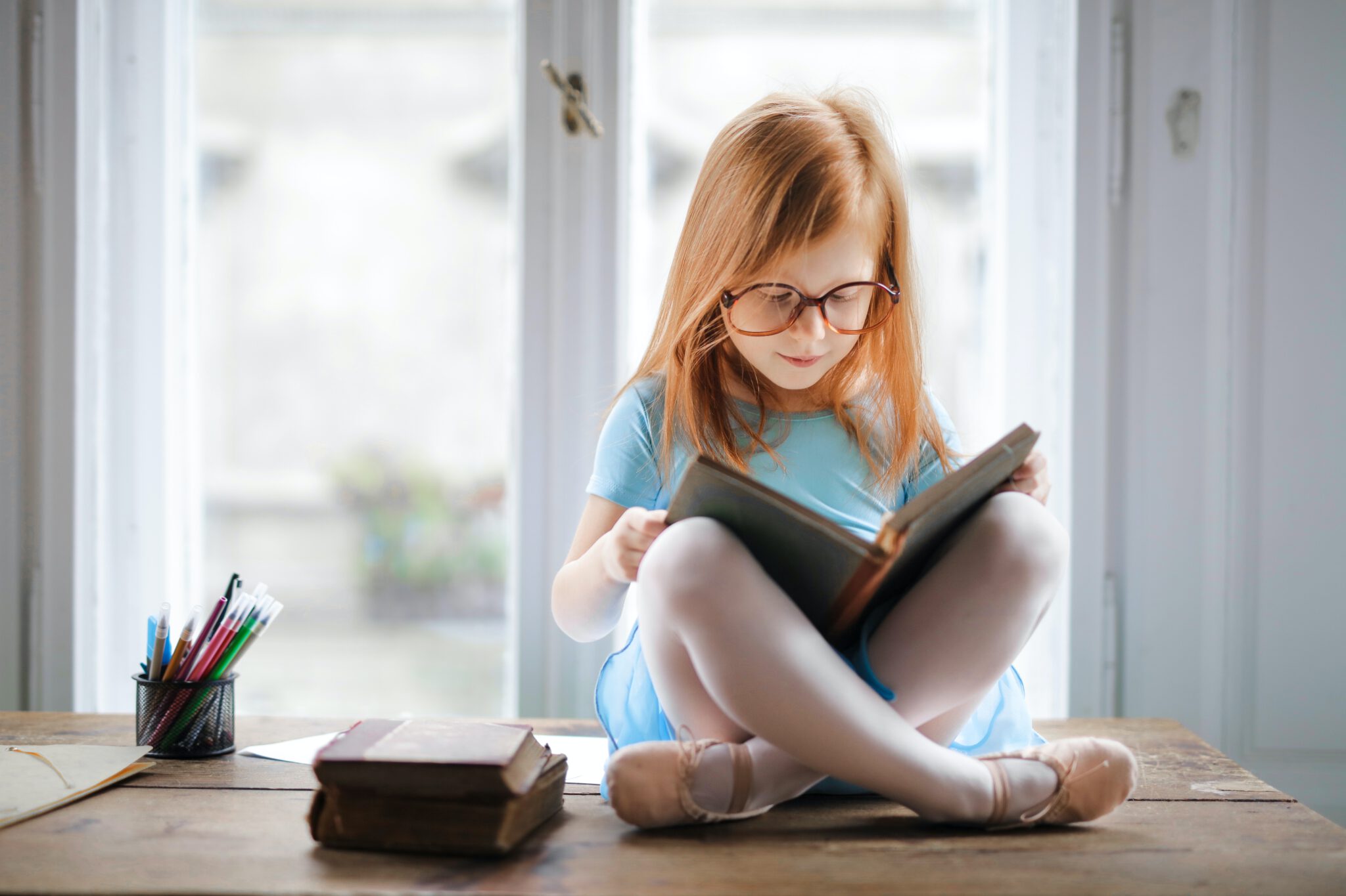 Roodharig meisje zit met een grote bril in kleermakerszit een boekje te lezen: een van de goede gewoonten die een kind kan aanleren